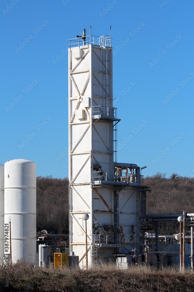 Tall old partially rusted white rectangle storage silo rising above row of brand new storage silos installed at the edge of modernized part of large oil refinery industrial complex surrounded with den Tall old partially rusted white rectangle storage silo rising above row of brand new storage silos installed at the edge of modernized part of large oil refinery industrial complex surrounded with den