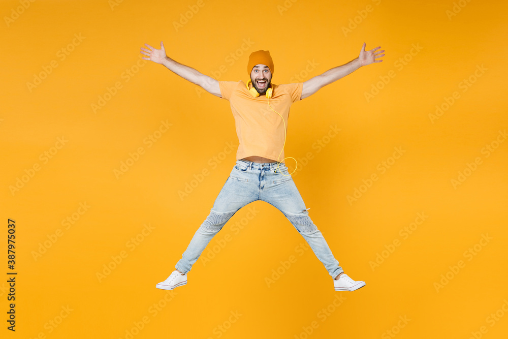 Full length of excited surprised young man 20s wearing basic casual t-shirt headphones hat jumping spreading hands and legs looking camera isolated on bright yellow colour background, studio portrait.
