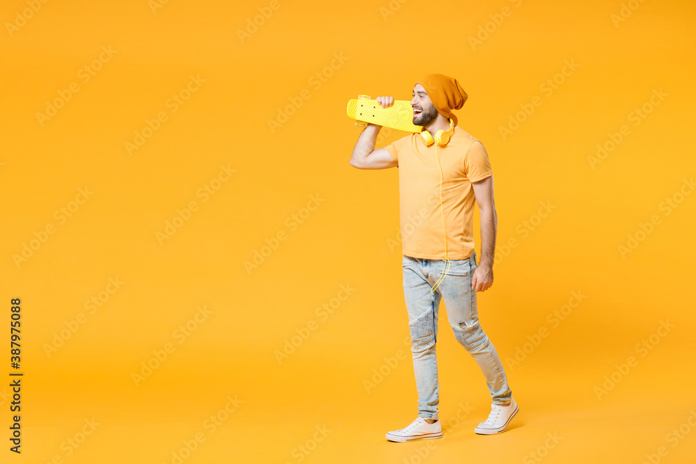 Full length side view of funny cheerful young man wearing basic casual t-shirt headphones hat walking going hold skateboard looking aside isolated on bright yellow colour background, studio portrait. Full length side view of funny cheerful young man wearing basic casual t-shirt headphones hat walking going hold skateboard looking aside isolated on bright yellow colour background, studio portrait.
