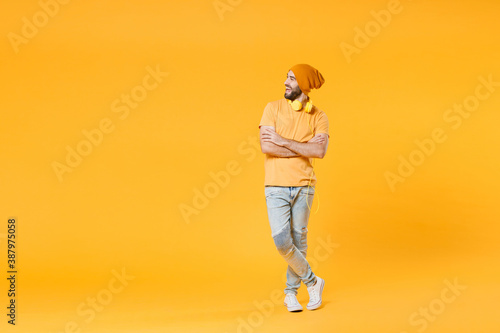 Full length of cheerful laughing young man 20s wearing basic casual t-shirt headphones hat standing holding hands crossed looking aside isolated on bright yellow colour background, studio portrait.