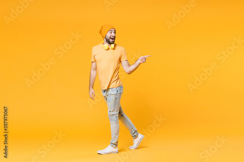 Full length side view of excited young man wearing basic casual t-shirt headphones hat standing pointing index finger aside on mock up copy space isolated on bright yellow background, studio portrait.
