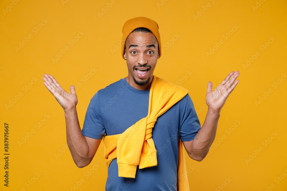 Excited surprised amazed young african american man 20s wearing basic casual blue t-shirt hat standing keeping mouth open spreading hands isolated on bright yellow colour background, studio portrait.