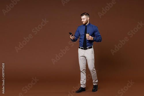 Full length successful young business man in blue shirt hold paper cup of coffee tea using mobile cell phone isolated on brown background studio portrait. Achievement career wealth business concept.