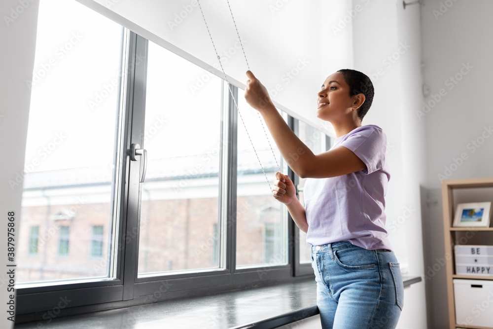 © Syda Productions - people and leisure concept - happy young african american woman opening window roller blinds at home
