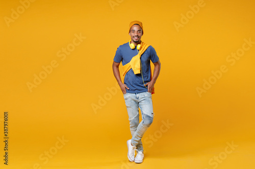 Full length of smiling handsome young african american man 20s wearing blue t-shirt hat standing holding hands in pockets looking camera isolated on bright yellow colour background, studio portrait.