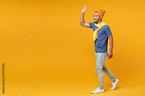 Full length side view smiling joyful young african american man 20s in blue t-shirt hat waving and greeting with hand as notices someone isolated on bright yellow colour background  studio portrait.