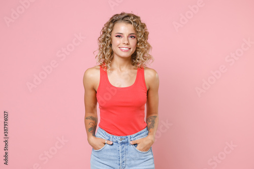 Smiling charming beautiful attractive young blonde woman 20s wearing basic casual tank top standing holding hands in pockets looking camera isolated on pastel pink colour background, studio portrait.