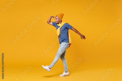 Full length side view of excited young african american man 20s in blue t-shirt hat standing holding hand at forehead looking far away distance isolated on bright yellow background, studio portrait.
