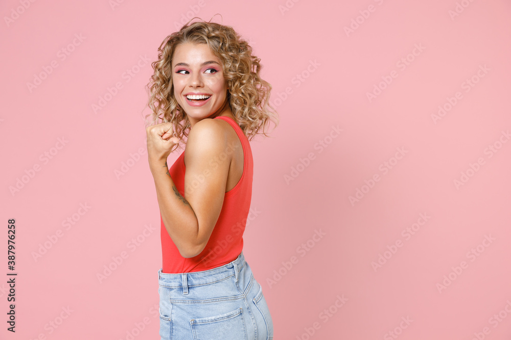 Side view of cheerful funny pretty young blonde woman wearing basic casual tank top standing pointing thumb aside on mock up copy space isolated on pastel pink colour wall background, studio portrait. Side view of cheerful funny pretty young blonde woman wearing basic casual tank top standing pointing thumb aside on mock up copy space isolated on pastel pink colour wall background, studio portrait.