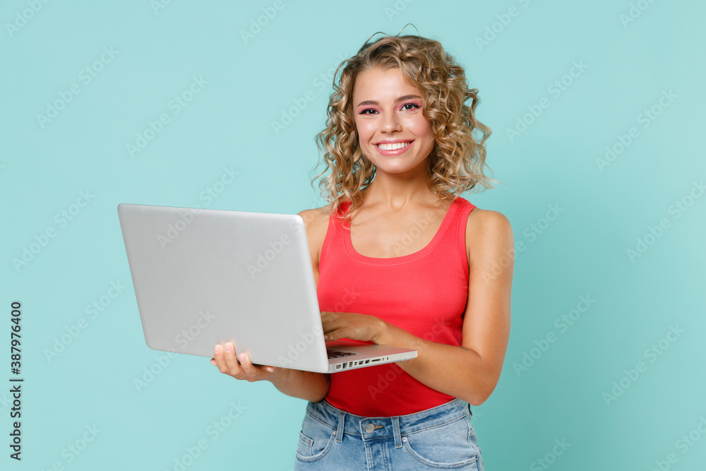 Smiling funny beautiful young blonde woman 20s wearing pink basic casual tank top standing working on laptop pc computer looking camera isolated on blue turquoise colour background studio portrait. Smiling funny beautiful young blonde woman 20s wearing pink basic casual tank top standing working on laptop pc computer looking camera isolated on blue turquoise colour background studio portrait.