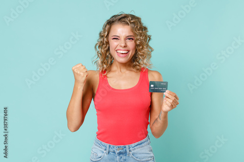 Joyful young blonde woman 20s wearing pink casual tank top standing hold in hand credit bank card doing winner gesture looking camera isolated on blue turquoise colour background studio portrait.