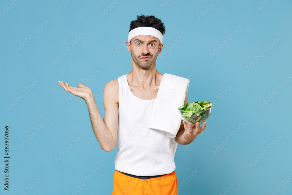 Confused sporty fitness man with thin skinny body sportsman in headband shirt shorts hold vegetable salad in bowl spreading hands isolated on blue background. Workout gym sport motivation concept. Confused sporty fitness man with thin skinny body sportsman in headband shirt shorts hold vegetable salad in bowl spreading hands isolated on blue background. Workout gym sport motivation concept.
