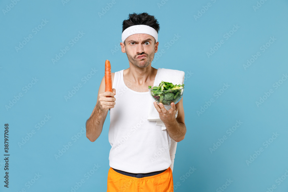 Confused young sporty fitness man with thin skinny body sportsman in headband shirt shorts hold vegetable salad in glass bowl carrot isolated on blue background. Workout gym sport motivation concept. Confused young sporty fitness man with thin skinny body sportsman in headband shirt shorts hold vegetable salad in glass bowl carrot isolated on blue background. Workout gym sport motivation concept.