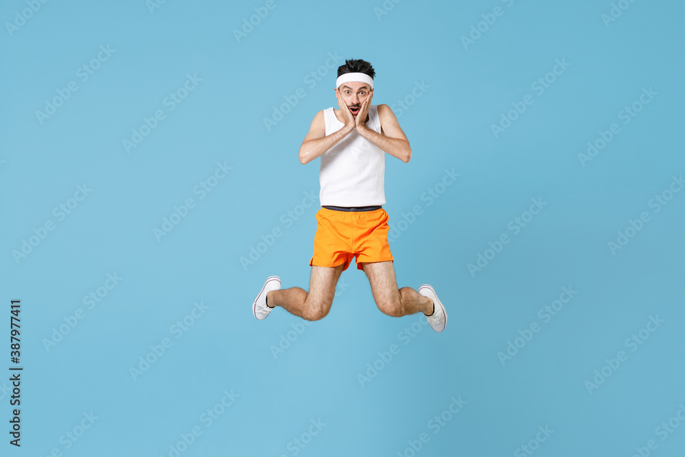 Full length portrait of shocked young fitness man with skinny body sportsman in headband shirt shorts jumping put hands on cheeks isolated on blue background. Workout gym sport motivation concept. Full length portrait of shocked young fitness man with skinny body sportsman in headband shirt shorts jumping put hands on cheeks isolated on blue background. Workout gym sport motivation concept.