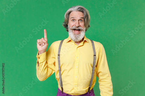 Excited elderly gray-haired mustache bearded man in casual yellow shirt suspenders hold index finger up with great new idea looking camera isolated on bright green colour background, studio portrait.