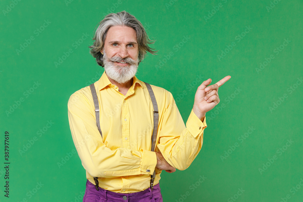 Smiling elderly gray-haired mustache bearded man wearing casual yellow shirt suspenders pointing index finger aside on mock up copy space isolated on bright green colour background studio portrait.