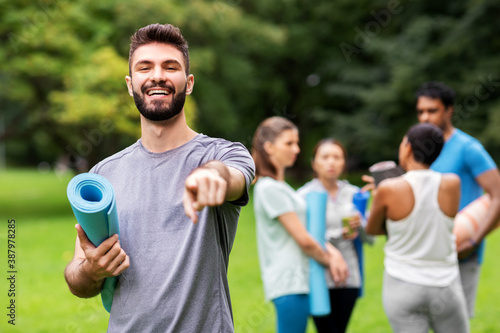 fitness, sport and healthy lifestyle concept - happy smiling young man with mat pointing finger to camera over group of people meeting for yoga class at summer park