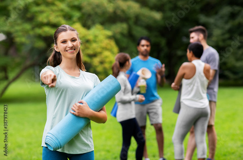 Fototapeta Naklejka Na Ścianę i Meble -  fitness, sport and healthy lifestyle concept - happy smiling young woman with mat pointing finger to camera over group of people meeting for yoga class at summer park