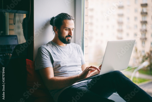 Obraz na płótnie Handsome attractive young dark-haired man working on laptop at home while sitting on windowsill with city view w salonie
