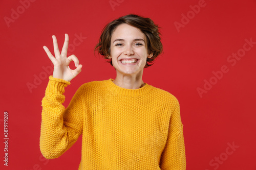 Smiling cheerful beautiful pretty young brunette woman 20s wearing basic casual yellow sweater standing showing OK gesture looking camera isolated on bright red colour background, studio portrait.