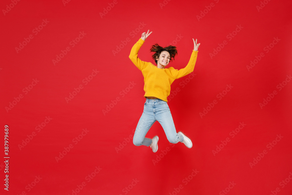 Full length of smiling cheerful funny young brunette woman 20s wearing basic casual yellow sweater jumping rising hands showing victory sign isolated on bright red colour background studio portrait. Full length of smiling cheerful funny young brunette woman 20s wearing basic casual yellow sweater jumping rising hands showing victory sign isolated on bright red colour background studio portrait.