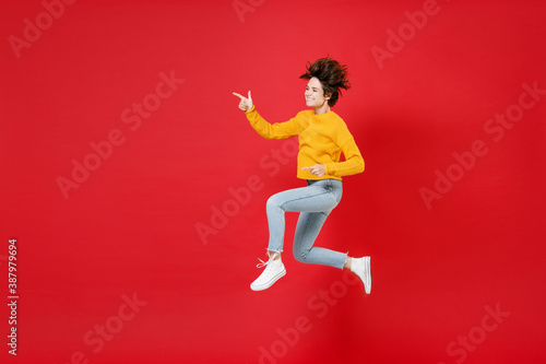 Full length side view of cheerful young brunette woman 20s in basic yellow sweater jumping pointing index fingers aside on mock up copy space isolated on bright red colour background studio portrait.