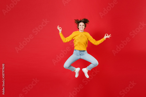 Full length of smiling young brunette woman 20s in basic yellow sweater jumping hold hands in yoga gesture relaxing meditating trying to calm down isolated on bright red background studio portrait.