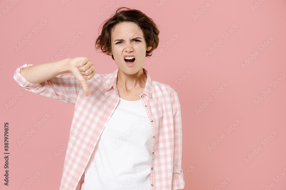 Dissatisfied displeased disgusted young brunette woman 20s wearing casual basic checkered shirt standing showing thumb down looking camera isolated on pastel pink colour background, studio portrait. Dissatisfied displeased disgusted young brunette woman 20s wearing casual basic checkered shirt standing showing thumb down looking camera isolated on pastel pink colour background, studio portrait.