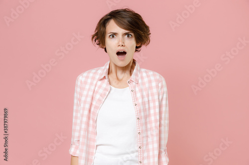 Shocked worried nervous irritated young brunette woman 20s wearing casual basic checkered shirt standing keeping mouth open looking camera isolated on pastel pink colour background, studio portrait.