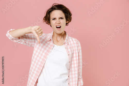 Dissatisfied displeased disgusted young brunette woman 20s wearing casual basic checkered shirt standing showing thumb down looking camera isolated on pastel pink colour background, studio portrait.