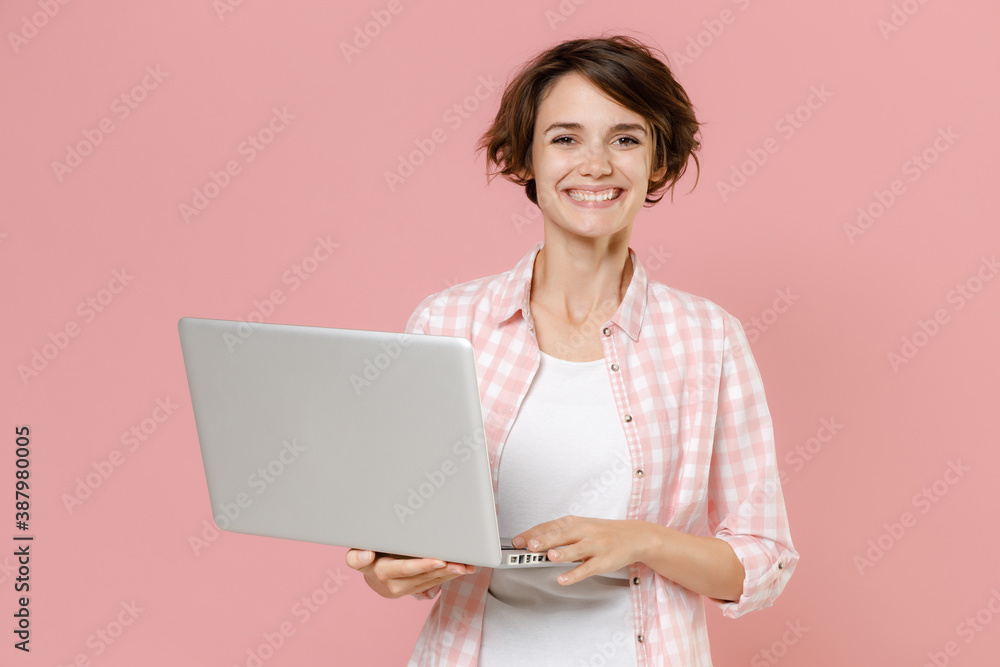 Smiling beautiful attractive young brunette woman 20s wearing casual checkered shirt standing working on laptop pc computer looking camera isolated on pastel pink colour background, studio portrait. Smiling beautiful attractive young brunette woman 20s wearing casual checkered shirt standing working on laptop pc computer looking camera isolated on pastel pink colour background, studio portrait.