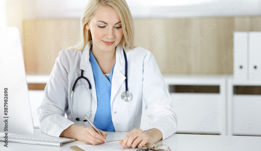 Woman-doctor at work while sitting at the desk in sunny clinic. Blonde cheerful physician filling up medication history record form Woman-doctor at work while sitting at the desk in sunny clinic. Blonde cheerful physician filling up medication history record form