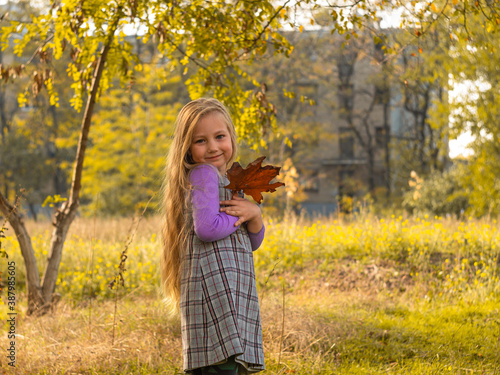 Beautiful girl with long blonde hair holds yellow leaves on nature background. Smiling child plays in autumn park outdoor on a sunny day. Cute happy kid in a plaid dress walking and dreaming alone.