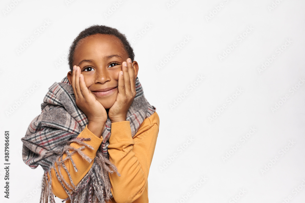 Studio shot of adorable dark skinned little boy in warm wrap holding hands on his cheeks and looking up with joyful smile, dreaming of something pleasant, feeling cold because of windy autumn day Studio shot of adorable dark skinned little boy in warm wrap holding hands on his cheeks and looking up with joyful smile, dreaming of something pleasant, feeling cold because of windy autumn day