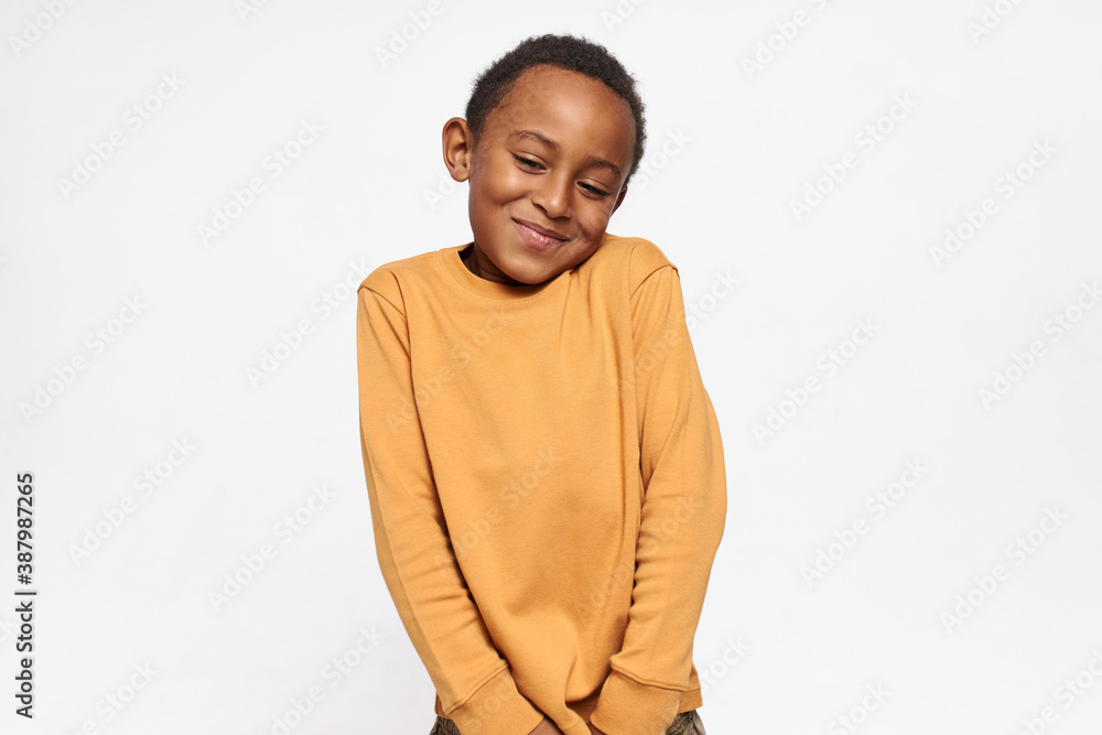 Horizontal shot of cute handsome dark skinned little boy shrugging shoulders feeling embarrassed with uncomfortable question, looking down with shy timid smile. Human emotions and body language Horizontal shot of cute handsome dark skinned little boy shrugging shoulders feeling embarrassed with uncomfortable question, looking down with shy timid smile. Human emotions and body language