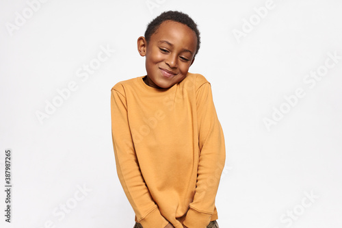 Cuadro en lienzo Horizontal shot of cute handsome dark skinned little boy shrugging shoulders feeling embarrassed with uncomfortable question, looking down with shy timid smile