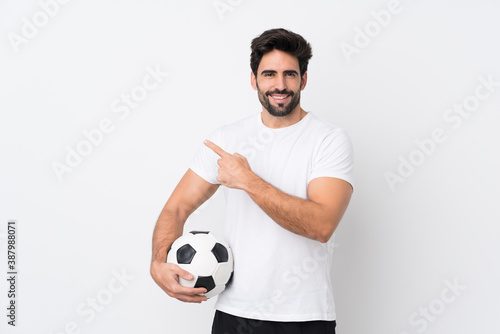 Young handsome man with beard over isolated white background with soccer ball and pointing to the lateral