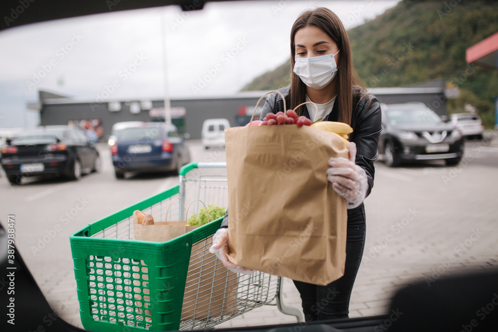 Over trunk view of young woman in medical mask in masks loading bags in  trunk after supermarket shopping. Quqrantine grocery shipping. Fresh fruits  for grandparents. Female with eco craft paket Stock Photo