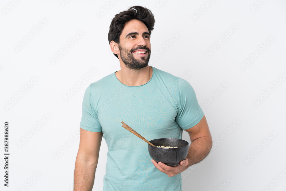 Young handsome man with beard over isolated white background looking up while smiling while holding a bowl of noodles with chopsticks Young handsome man with beard over isolated white background looking up while smiling while holding a bowl of noodles with chopsticks