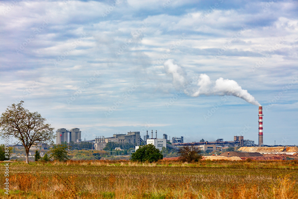 The production complex of a cement plant on the horizon behind an agricultural field. The production complex of a cement plant on the horizon behind an agricultural field.