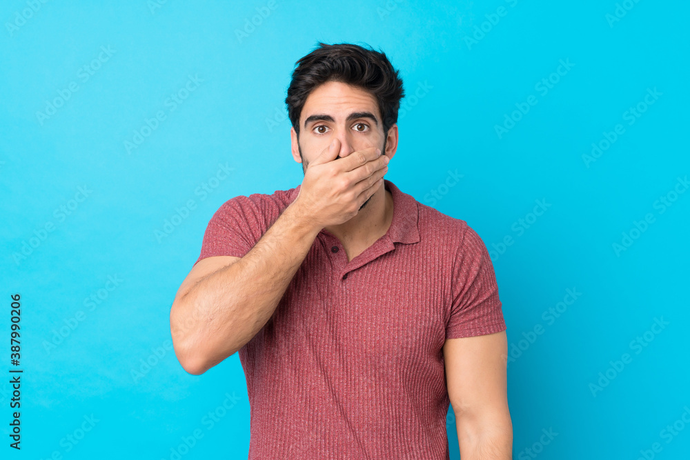 Young handsome man with beard over isolated blue background covering mouth with hands Young handsome man with beard over isolated blue background covering mouth with hands