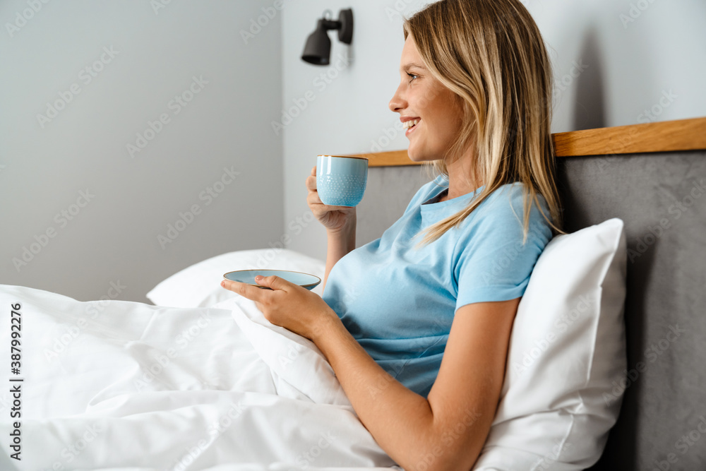 Happy young woman laying in bed, drinking coffee morning