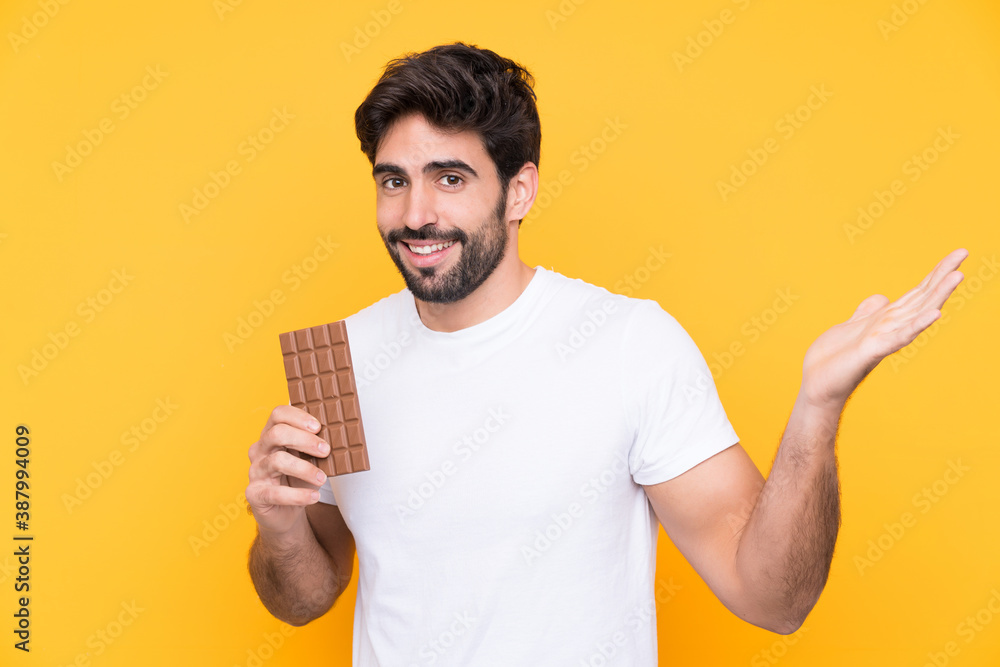 Young handsome man with beard over isolated yellow background taking a chocolate tablet and surprised Young handsome man with beard over isolated yellow background taking a chocolate tablet and surprised