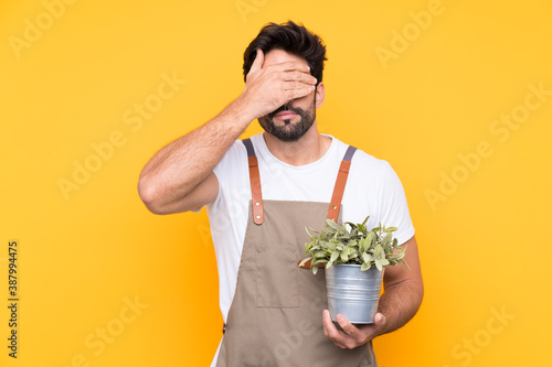Gardener man with beard over isolated yellow background covering eyes by hands