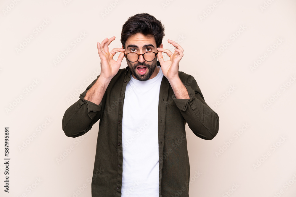 Young handsome man with beard over isolated background with glasses and surprised Young handsome man with beard over isolated background with glasses and surprised