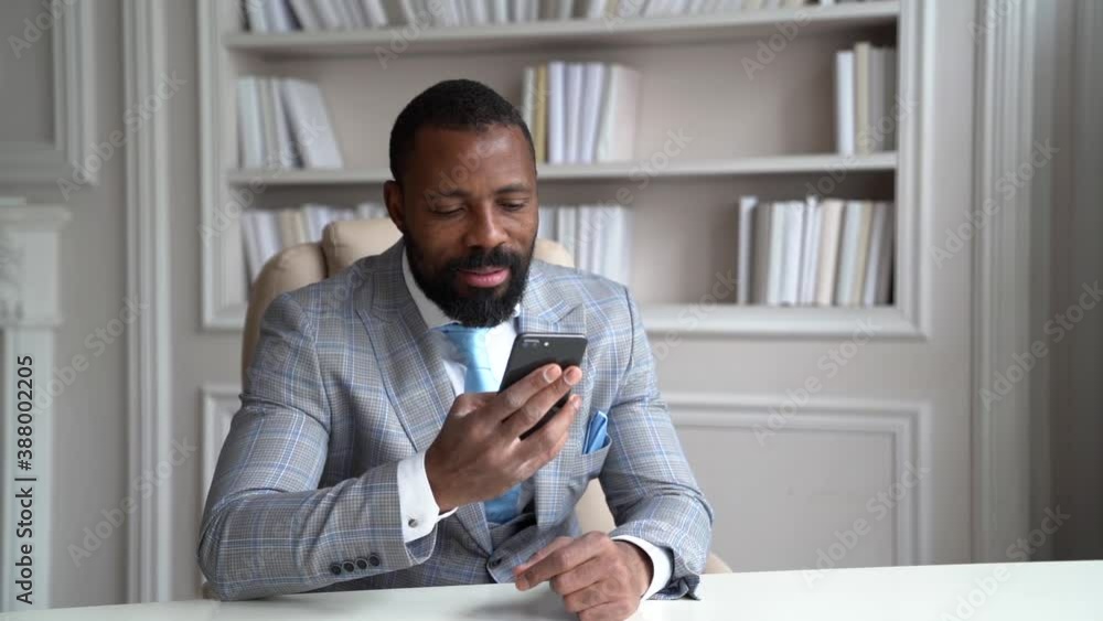 Portrait of a young successful African businessman in a suit sitting at his office Desk and reading a message on his mobile phone Portrait of a young successful African businessman in a suit sitting at his office Desk and reading a message on his mobile phone