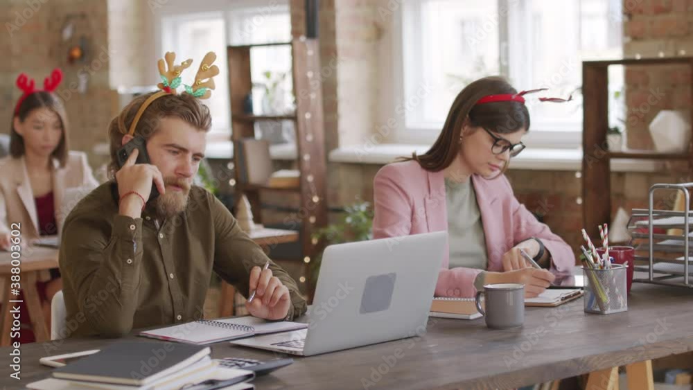 PAN shot of business colleagues wearing festive headbands sitting at desks in open plan office and working on Christmas Eve or New Years Day PAN shot of business colleagues wearing festive headbands sitting at desks in open plan office and working on Christmas Eve or New Years Day
