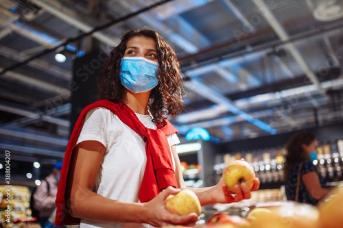 Young woman picks up some vegitables and fruits at a supermarket wearing a medical sterile mask during the coronavirus pandemic quarantine. Healthcare, stay home and sanitizing concept.