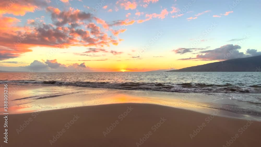 Wide shot of gentle waves rolling to shore in a tropical island sunset with sandy foreground and a distant island in the distance with fluffy pink clouds set against a light blue sky Wide shot of gentle waves rolling to shore in a tropical island sunset with sandy foreground and a distant island in the distance with fluffy pink clouds set against a light blue sky