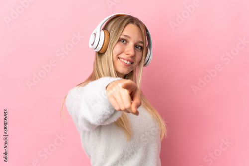 Young blonde woman isolated on pink background listening music and pointing to the front